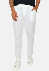 Pantaloni bianchi in lino con vita regolabile, gambe affusolate e tasche laterali, abbinati a sneakers bianche su uno sfondo neutro.