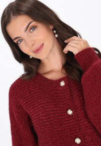 Bourgogne stickad cardigan med en texturerad yta, rund halsringning och tre pärlprydda knappar fram. Långa ärmar.
