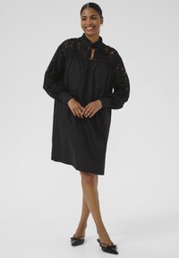Abito nero con maniche lunghe in pizzo, scollatura a chiave e colletto arricciato. Presenta una silhouette fluida e un design minimalista.