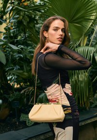 Beżowa skórzana torebka z złotymi akcentami na górze, miękka, zaokrąglona forma, trzymana przez osobę w dopasowanej wzorzystej sukience na tle zieleni.