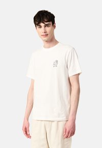 Kermaati puuvillapaita, jossa on minimalistinen musta graafinen kuva kädestä ja teksti "LA DOLCE VITA" vasemmassa rinnassa. Lyhyet hihat, rento malli.