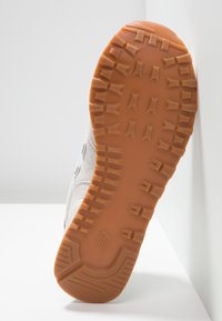 Semelle de chaussure en caoutchouc brun clair avec un motif de bande de roulement profond et multidirectionnel, montrant partiellement la partie supérieure grise claire de la chaussure.