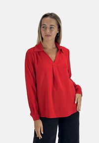 Camicia rossa a maniche lunghe con scollatura a V e colletto, realizzata in tessuto testurizzato e dalla vestibilità rilassata. Indossata con pantaloni neri.