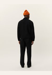 Chaqueta negra de textura con cuello, llevada sobre pantalones negros. Un gorro de lana naranja añade un toque de color en contraste.