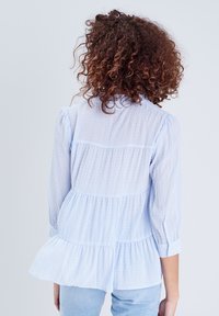 Blouse rayée bleu clair avec ourlet froncé et manches trois-quarts. Fabriquée en tissu léger, avec une coupe ample et un design en couches.
