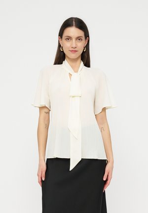 Hosszú barna hajú nő fehér, rövid ujjú blúzt visel nyakkendővel és fekete szoknyát, egy egyszínű háttér előtt áll.