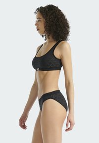 Žena s kudrnatými vlasy stojí bokem, na sobě má černou sportovní podprsenku se vzorem a odpovídající tanga, na světlém pozadí.