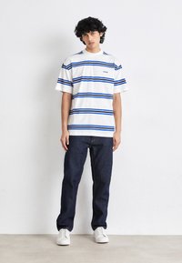 T-shirt bianca a maniche corte con righe orizzontali blu e logo "HUGO" sul petto, abbinata a jeans blu scuro e sneakers bianche.