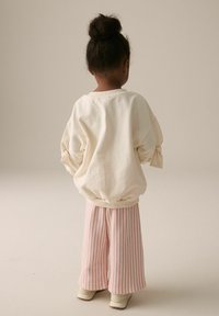 Helles beiges Sweatshirt mit gerafftem Bund und Ballonärmeln, kombiniert mit rosa-weiß gestreiften, weiten Hosen, getragen mit hellen Sneakers.