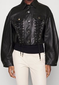 Svart skinnjacka i kort modell med knäppning framtill, snörningsdetaljer och bronsfärgade metalldelar, med ett svart elastiskt midjeband.