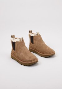 Botas de tobillo de ante marrón con un suave forro de piel de oveja blanca, paneles laterales elásticos y una suela de goma texturizada. Presentan costuras de contraste.
