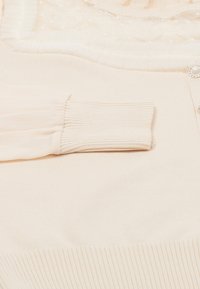 Top color beige chiaro con tessuto a costine, maniche a sbuffo trasparenti, design a balze sovrapposte e dettaglio di bottoni decorativi.