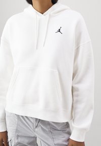 Sudadera blanca con capucha, hecha de una tela suave, que presenta un bolsillo frontal y un logo de Jordan negro en el pecho. Diseño de corte holgado con puños acanalados.