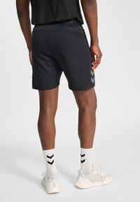 Schwarze Sportshorts mit glatter Textur, ausgestattet mit einem breiten Taillenbund und einem grauen Chevron-Muster an der Seite. Weiße Turnschuhe und Socken.