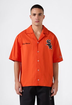 Hombre con una camisa de manga corta de color naranja brillante con el logo y texto de Chicago White Sox, de pie frente a un fondo blanco liso.