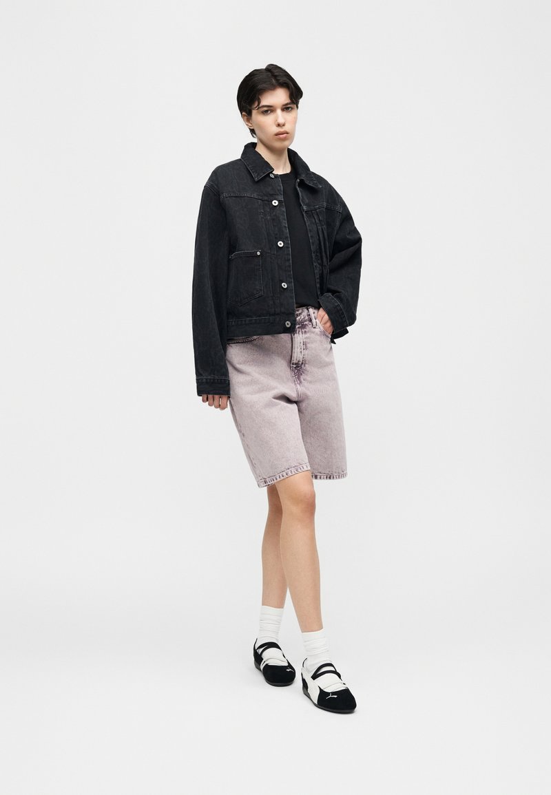 Giovane che indossa una giacca di denim nera, top nero, pantaloncini di denim viola chiaro, calzini bianchi e scarpe bianco e nero, in piedi contro uno sfondo uniforme.