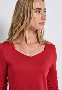 Rotes langärmliges Oberteil mit einem V-Ausschnitt, aus weichem Stoff gefertigt. Hat eng anliegende Ärmel und eine glatte Textur. Ohrringe sind sichtbar.