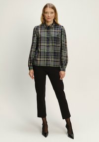 Košulja dugih rukava u kariranoj kombinaciji tamnozelene, mornarskoplave i bež boje, s dugmadi sprijeda i klasičnim kragnom, uparena s crnim hlačama i cipelama na visoku petu.