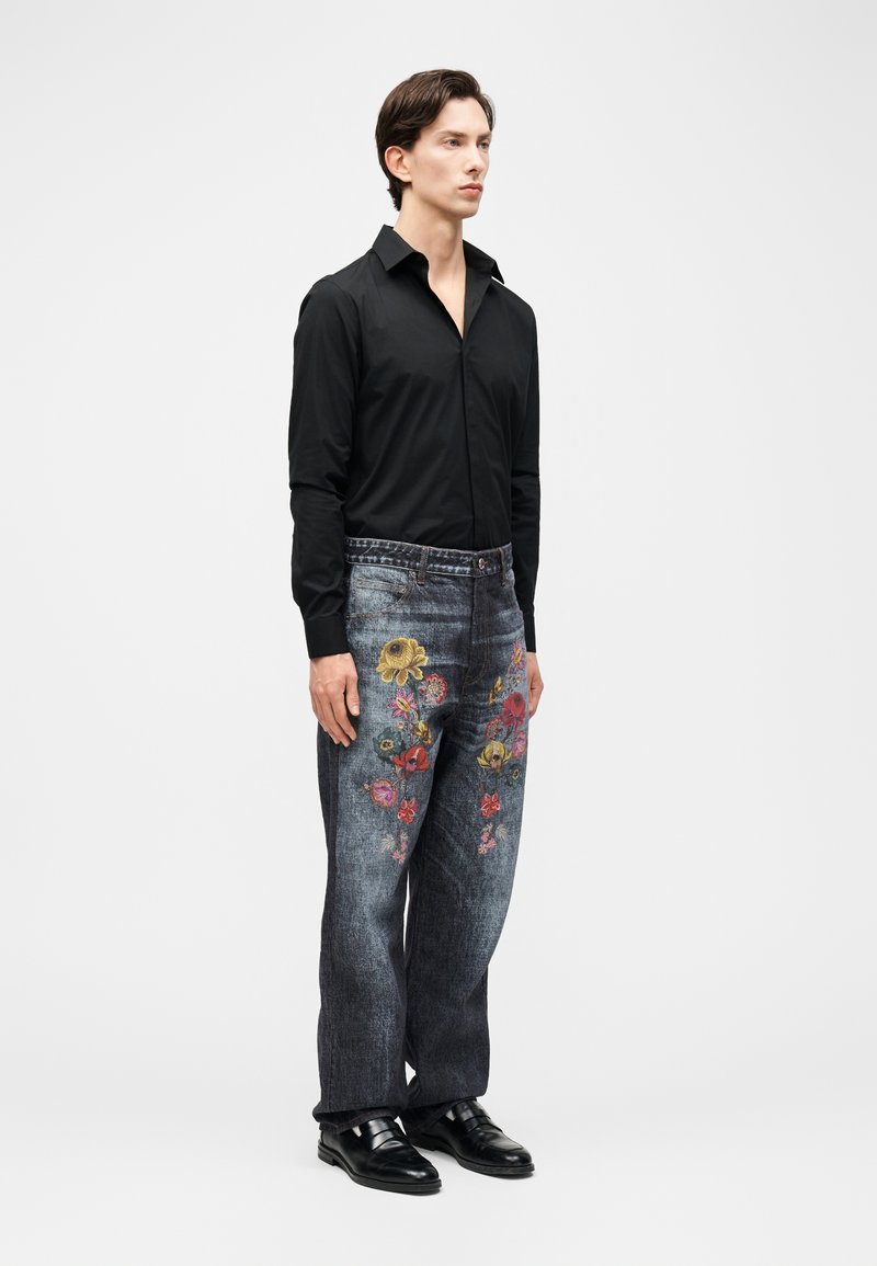 Homme portant une chemise noire à manches longues, un jean foncé avec une broderie florale colorée, et des chaussures en cuir noir, debout devant un fond blanc.