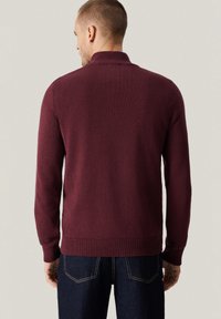 Bordowy sweter z dzianiny z prążkowanym kołnierzem i mankietami, o gładkiej fakturze i dopasowanym kroju, zestawiony z ciemnymi dżinsami.