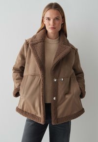 Bruine shearling gevoerde jas met een kraag, zijzakken en knoopdetails. Draag over een beige trui en gecombineerde met donkere spijkerbroeken.