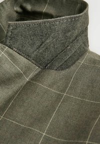 Gros plan sur un tissu en laine verte avec un motif à fines carreaux blancs en damier et un col plié avec des coutures visibles.