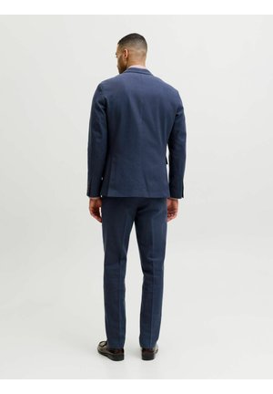 Férfi áll háttal, sötétkék öltönyt visel hozzáillő nadrággal és fekete elegáns cipőkkel, egyszerű világos háttér előtt.