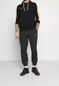 Mężczyzna w czarnej bluzie Nike z złotymi paskami na rękawach, czarnych spodniach dresowych Nike, białych skarpetkach i czarno-czerwonych butach Nike.