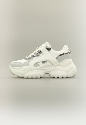 Baskets blanches épaisses avec des accents à motif de peau de serpent, semelle épaisse texturée, et bride Velcro sur les lacets sur fond neutre.