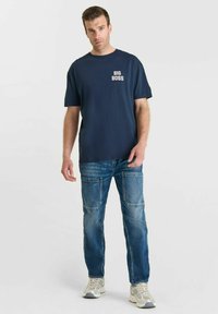 Man in navy "BIG BOSS" t-shirt, blauwe jeans met grote voorzakken en witte sneakers, staand tegen een effen witte achtergrond.
