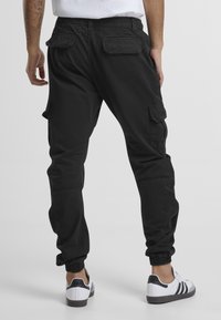 Zwarte cargobroek met elastische boorden en meerdere zakken. Gemaakt van stof met een gladde textuur. Draagt onder witte en zwarte sportschoenen.