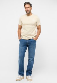 Beżowa bawełniana koszulka z okrągłym dekoltem i białym logo, w połączeniu z niebieskimi dżinsami. Model nosi jasne trampki.
