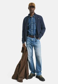 Man som bär en rutiga keps, marinblå zip-jacka över en denimskjorta, ljusblå jeans, brunt bälte, håller en brun kappa och har på sig vandringskängor.