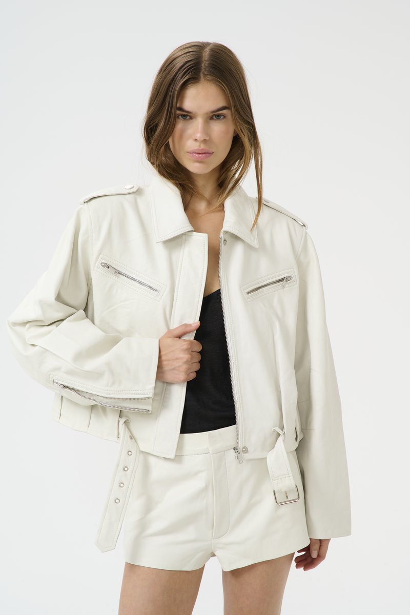 White leather cropped jacket with shoulder epaulettes, two zippered front pockets, and belted detail. Paired with white leather shorts.