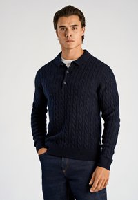 Marineblauer Pullover mit Zopfmuster, Kragen und drei Knöpfen, strukturiertem Design sowie gerippten Bündchen und Saum. Getragen zu dunklen Jeans.