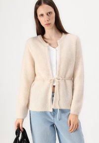 Krémový pletený cardigan s páskem na zavazování, kulatým výstřihem a žebrovanými manžetami, v kombinaci s bílou košilí na knoflíky a modrými džíny.