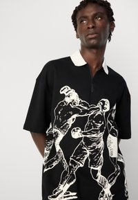 Young man with dark skin and curly hair wearing black polo shirt with white collar and graphic of two boxers fighting.