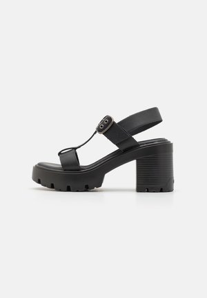 Sandalias negras de tacón con plataforma gruesa, que presentan una textura suave, correa ajustable en el tobillo con un botón circular y suela con relieve.