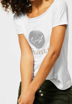 Person med krøllet hår iført en hvid T-shirt med en grå cirkel og teksten "trust yourself,", kombineret med mørkegrønne bukser med dekorative knapper.