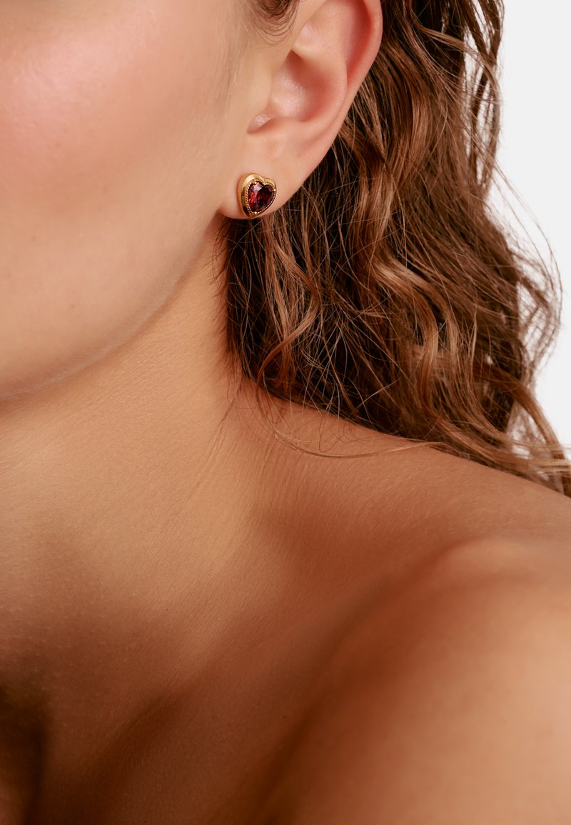 Gros plan de l’oreille d’une personne portant une boucle d’oreille en forme de cœur en or avec une pierre rouge, avec des cheveux bouclés bruns visibles.