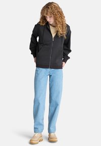 Schwarze Kapuzenjacke mit Reißverschluss, Kordelzug und gerippten Bündchen; über einem beigen Shirt getragen und mit hellblauen Jeans kombiniert.