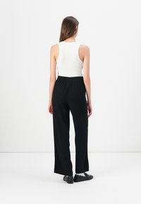 Zwarte wijde broek met een elastische tailleband, gecombineerd met een aansluitend wit geribbeld tanktop. De achtergrond is eenvoudig wit.