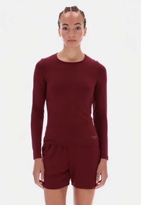 Modelka ubrana w dopasowaną bordową bluzkę z długim rękawem oraz pasujące szorty, stojąca prosto na jednolitym jasnym tle.