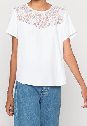 Witte blouse met korte mouwen, een bloemrijk kanten inzetstuk en een ronde halslijn, gedragen met blauwe spijkerbroek, afgebeeld van midden van de torso tot de heupen.