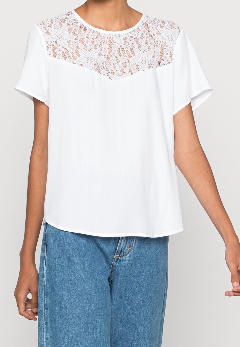 Blouse blanche à manches courtes avec empiècement en dentelle florale et encolure ronde, portée avec un jean denim bleu, vue du milieu du torse jusqu'aux hanches.