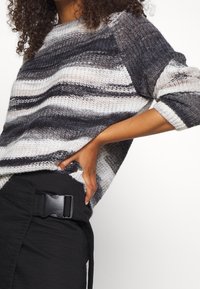 Frau, die einen gestreiften grau-weißen Strickpullover mit hochgekrempelten Ärmeln und schwarze Hosen mit einem großen Schnallen-Gürtel trägt.