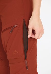 Ruosteenväriset housut, jotka on valmistettu teksturoidusta kankaasta. Housuissa on vetoketjulliset taskut ja avattavasta taskusta näkyvä verkkovuori. Sivulla on logoyksityiskohta.