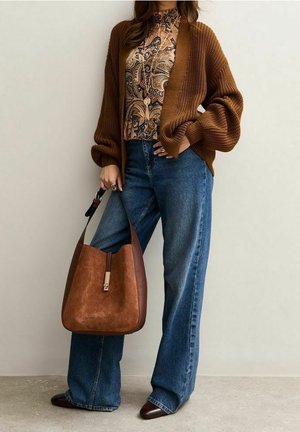 Femme portant un cardigan en maille marron, un chemisier à motifs, un jean bleu à jambes larges, tenant un grand sac à main marron en daim et cuir.