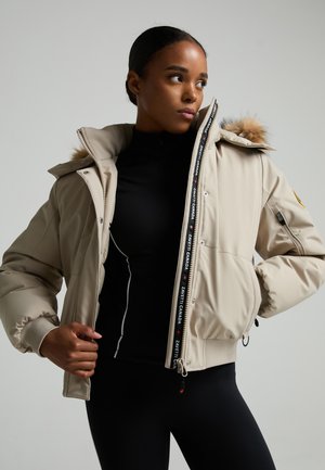 Vrouw draagt een beige donsjas met bontkap over een zwarte outfit, houdt de jas open en kijkt opzij tegen een effen achtergrond.