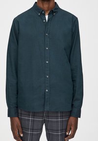 Donkerblauwe button-up shirt, gemaakt van zachte stof. Beschikt over een kraag, knopen aan de voorkant en lange mouwen. Draagt over een grijze geruite broek.